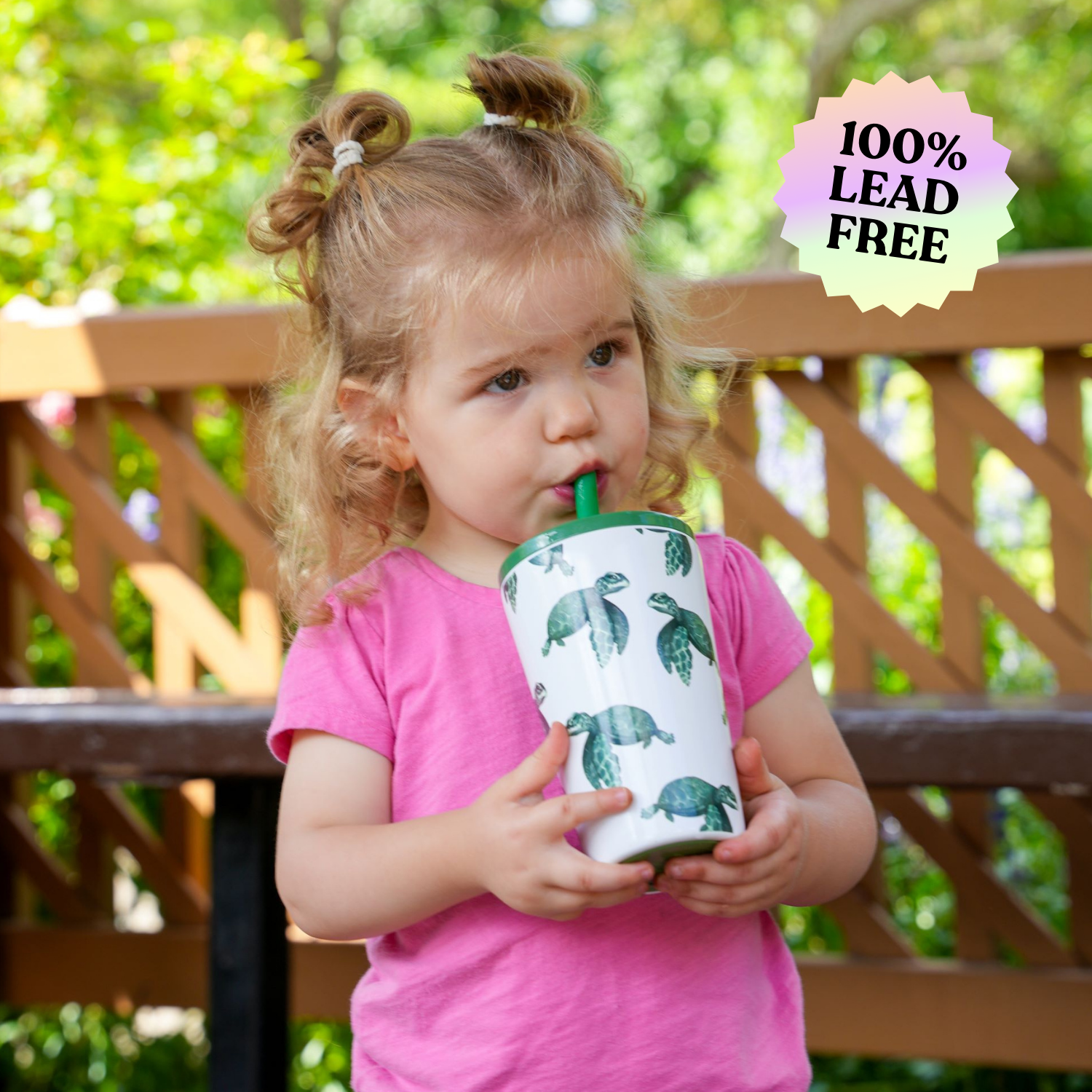 Young girl drinking from a 14 oz stainless steel cup with a sea turtle design, featuring a built-in soft silicone straw, outdoors on a sunny day with a wooden fence in the background. The cup is labeled as 100% lead-free.